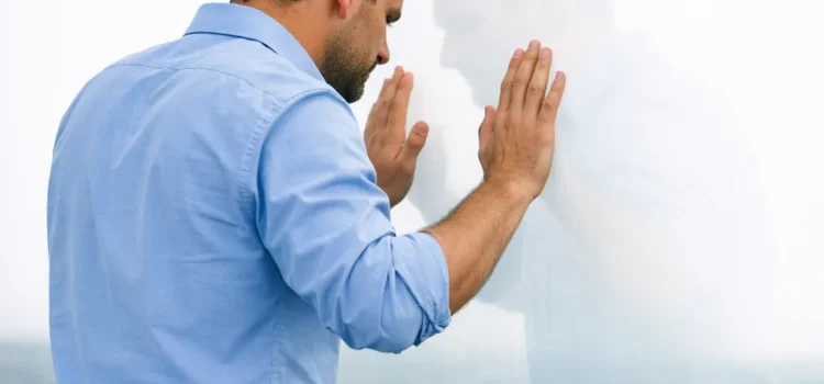 Homme de dos en chemise bleue face à une barrière invisible, les mains posées contre une surface transparente, symbolisant les croyances limitantes.